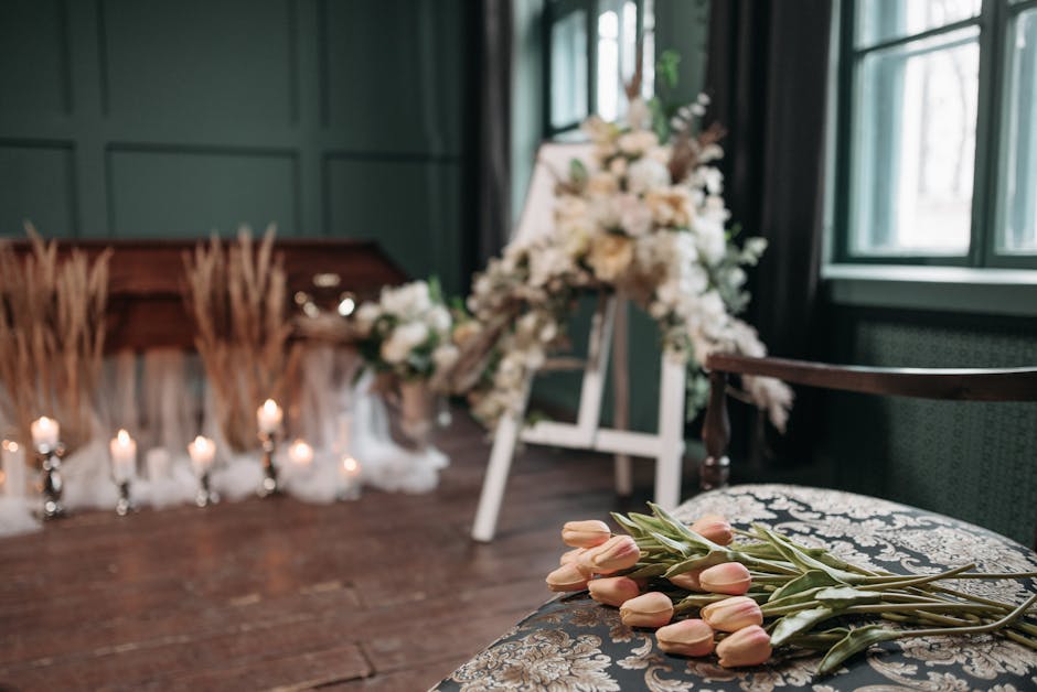 A collection of pale pink tulips with long green stems and leaves resting on an ornate fabric-covered chair. In the background, there is a floral arrangement composed of white roses, peonies, and gree