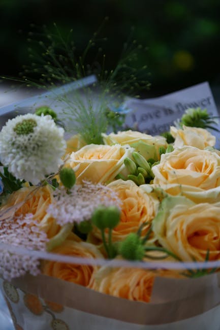 A close-up of a floral bouquet featuring soft peach-colored roses with gently unfurled petals, complemented by small white pom-pom chrysanthemums and delicate green buds. The arrangement is wrapped in
