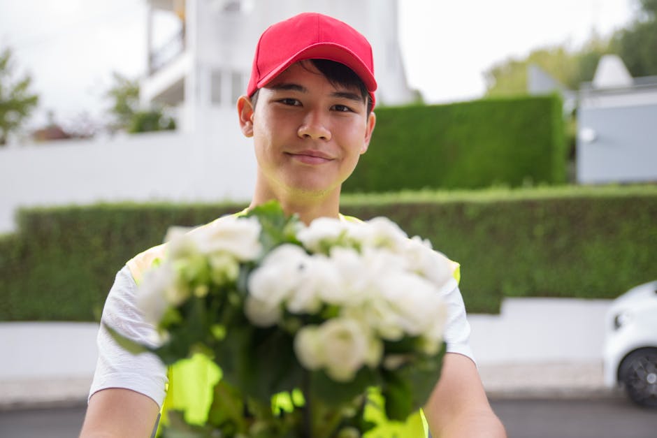 A delivery person wearing a bright yellow high-visibility vest and a red cap presents a bouquet of fresh white flowers with green foliage, wrapped in clear plastic, outdoors on a city street. The indi