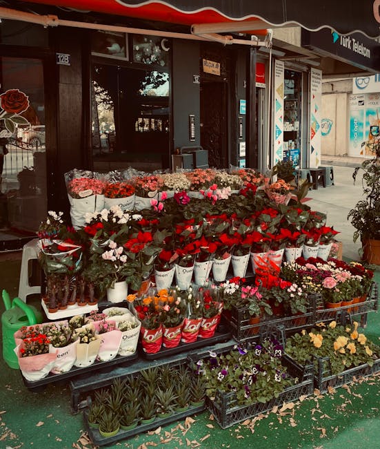 A vibrant outdoor display of fresh flowers arranged on multi-tiered black metal racks in front of a shop with a dark entrance. The arrangement features a variety of colorful blossoms including red ros