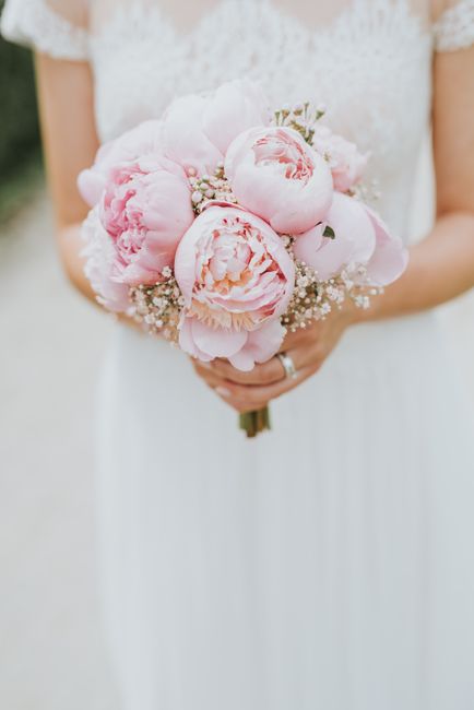 A bride holding a bouquet arrangement featuring large, soft pink peonies with delicate, ruffled petals and hints of cream, accented with small white baby's breath flowers. The bouquet is wrapped with 